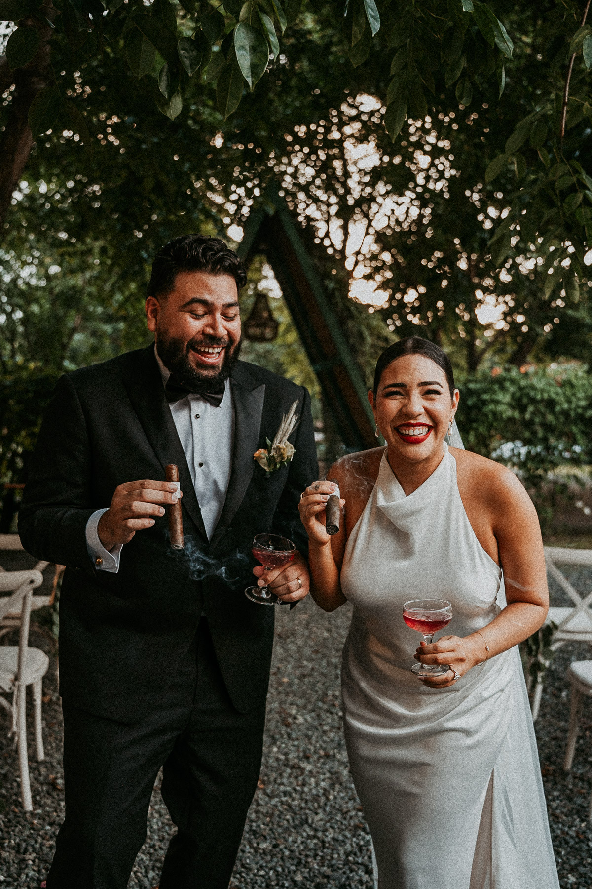 Candid photo of a newlywed couple laughing and smoking artisanal cigars at their alternative mountain wedding.