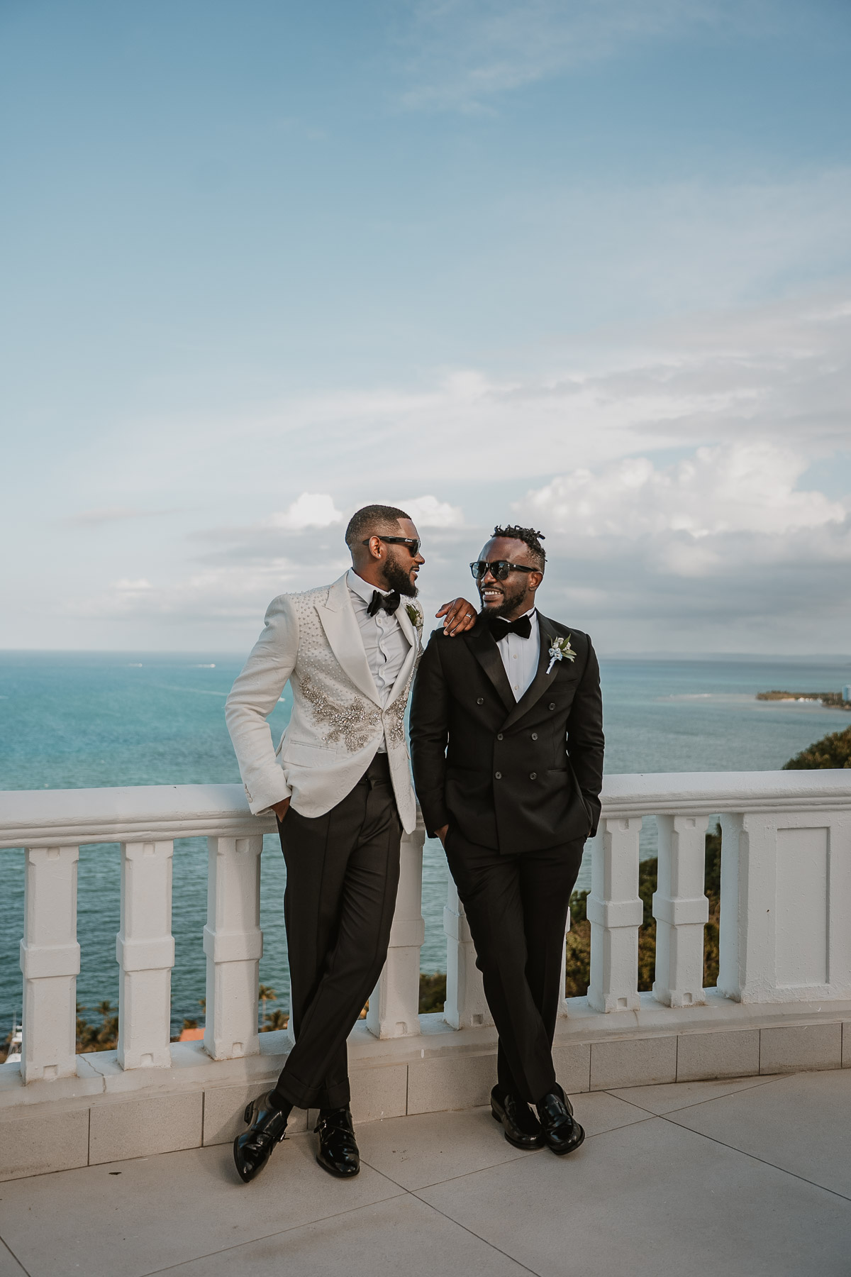 Two grooms standing together on balcony overlooking Fajardo coastline