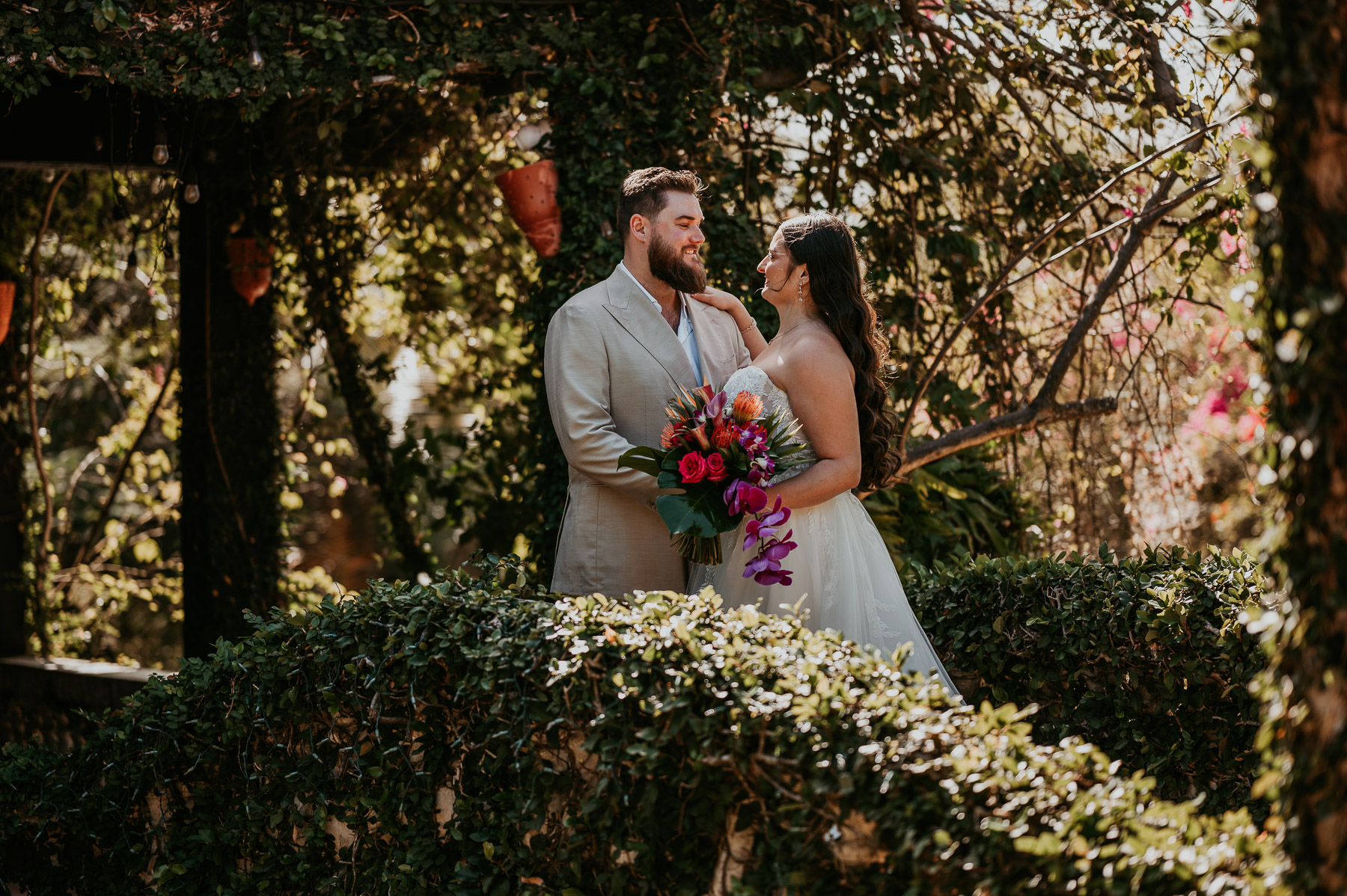Wide shot of bride and groom embracing beneath the tropical greenery