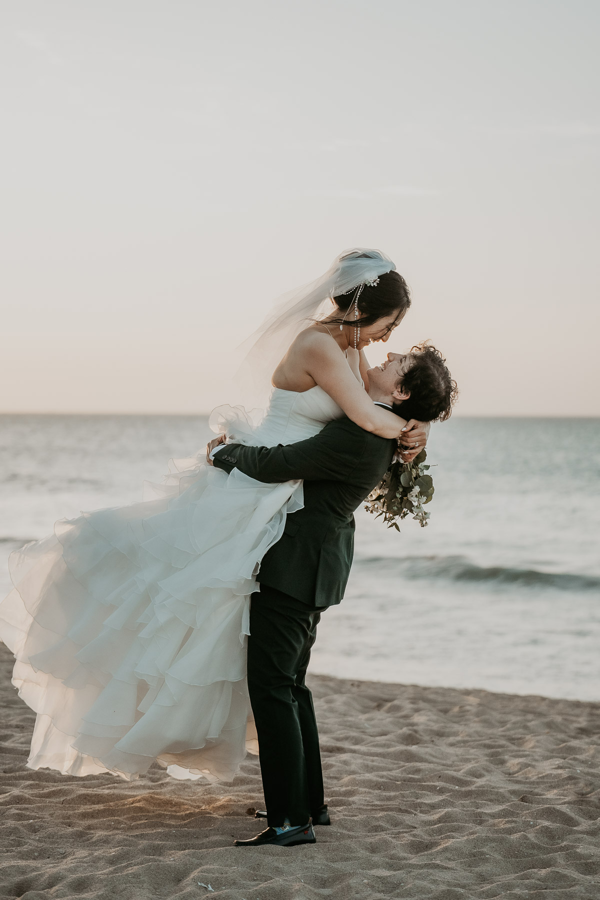 An unposed, tender moment between a newlywed couple standing near the ocean waves.