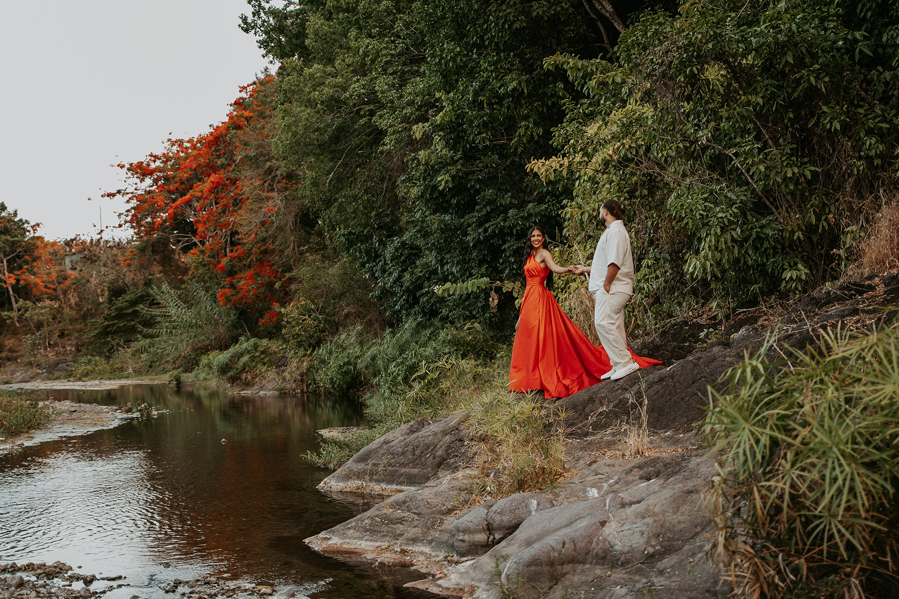 Bride in a striking orange-red dress posing beside the river during a Puerto Rico adventure session